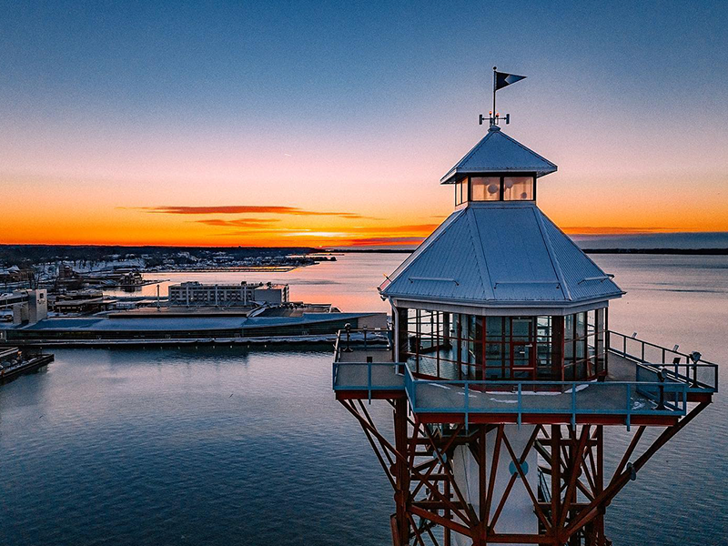 A lighthouse with a white roof stands over calm water at sunset, with orange and blue hues in the sky and distant buildings along the shoreline.