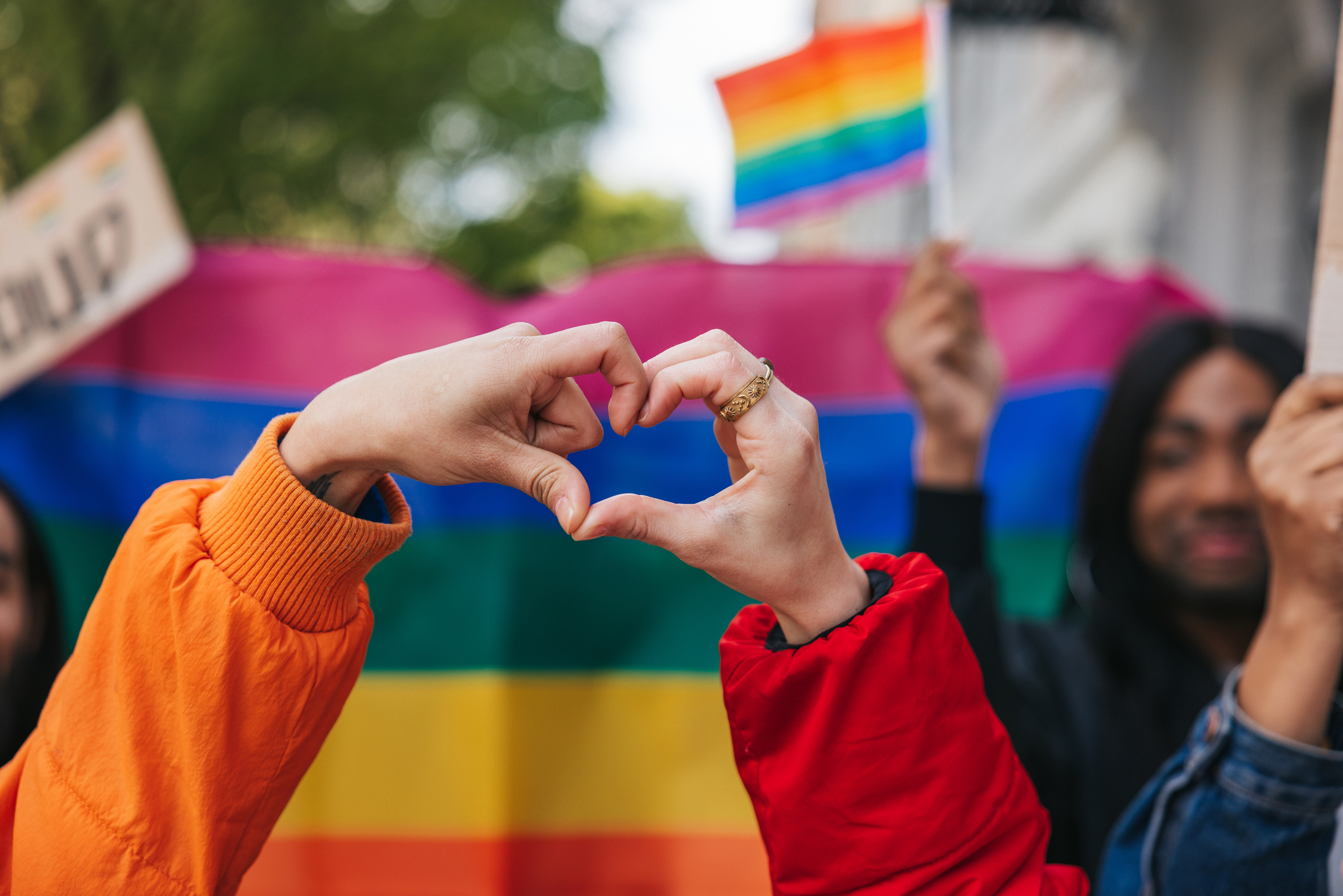 Genderqueer and non-binary friends making a heart shape against a rainbow flag