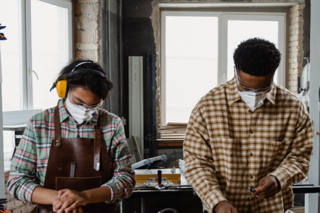 Two people wearing protective glasses and masks work with tools in a workshop. One person also wears ear protection and an apron; both are dressed in plaid shirts and stand near a window with equipment on the table.