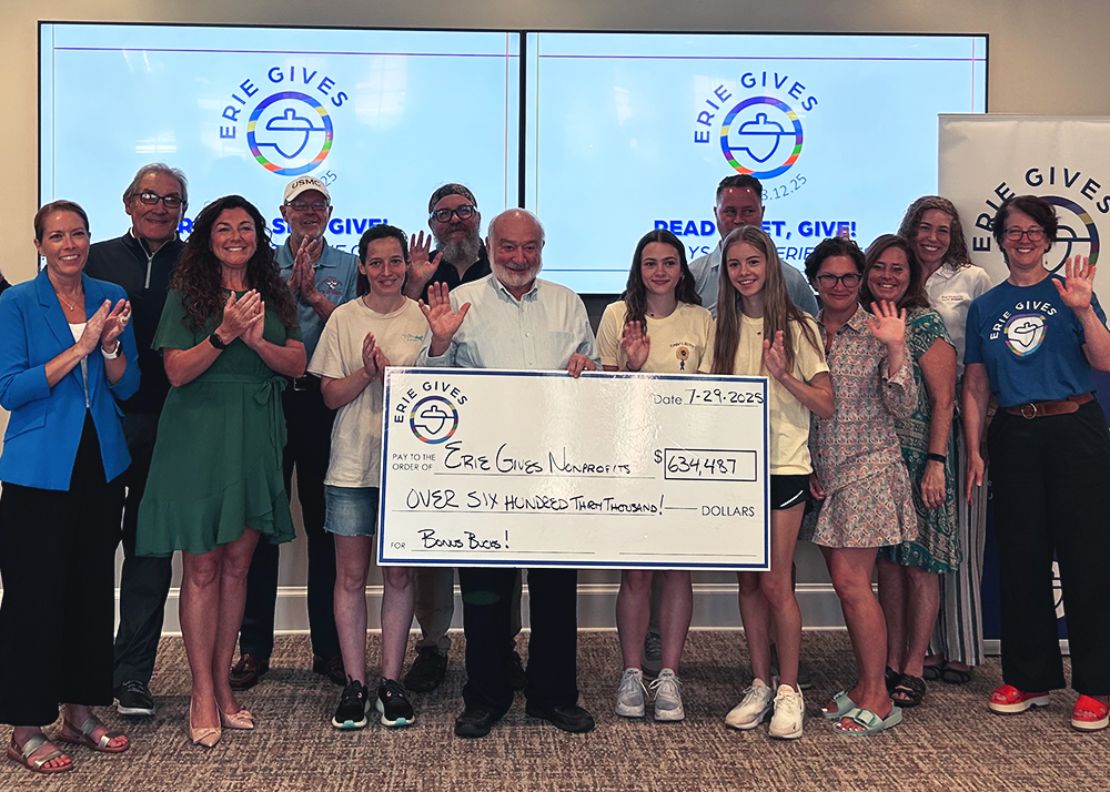 A group of people standing and smiling indoors, holding an oversized check for $634,481 made out to Erie Gives Nonprofits. Two large screens behind them display the Erie Gives logo.