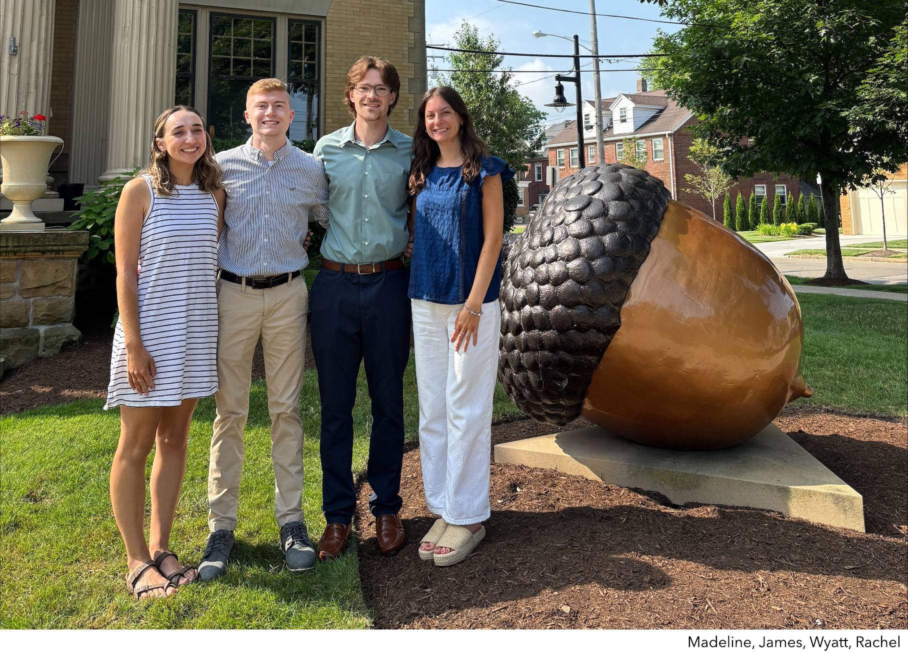 Four people stand smiling together outdoors next to a large acorn sculpture on a lawn. Two women and two men are casually dressed, with buildings and trees in the background.