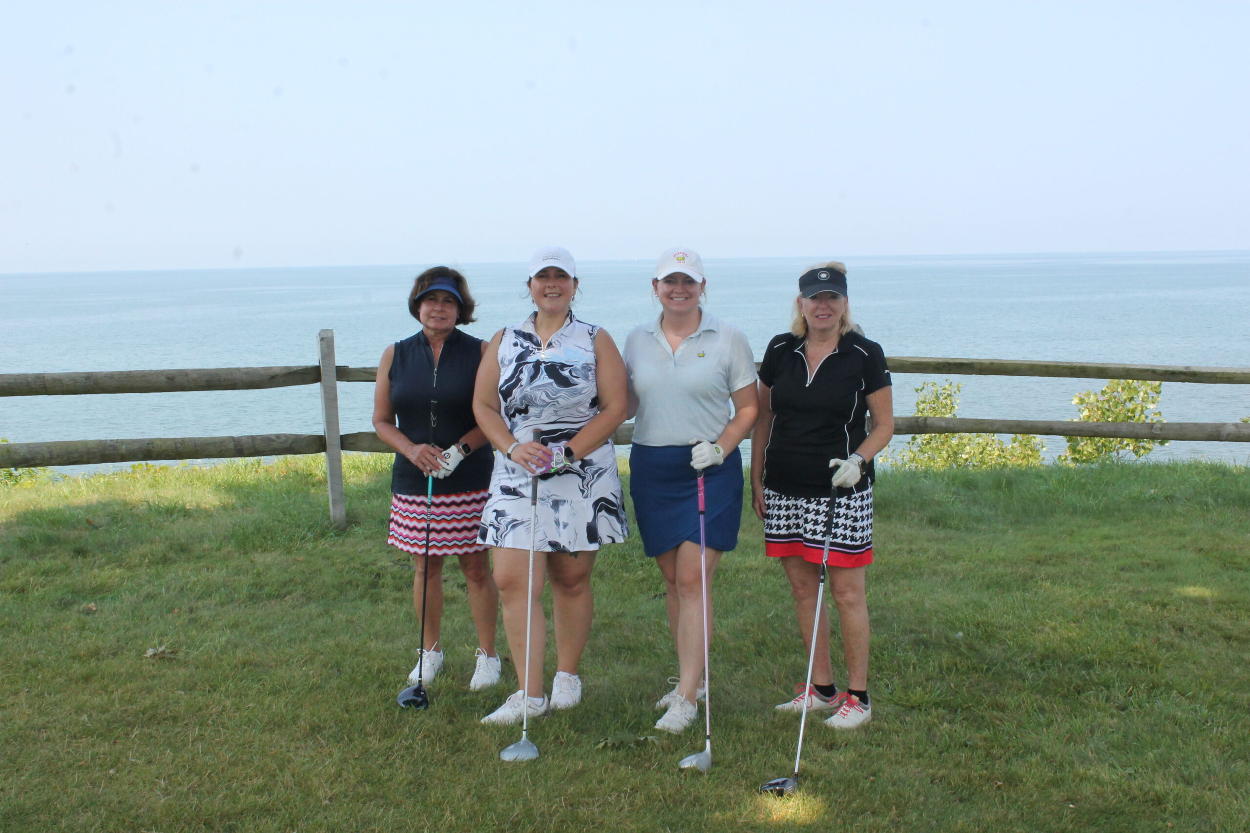 Four women in golf attire stand on grass holding golf clubs, smiling at the camera. Behind them is a wooden fence and a large body of water under a clear sky.