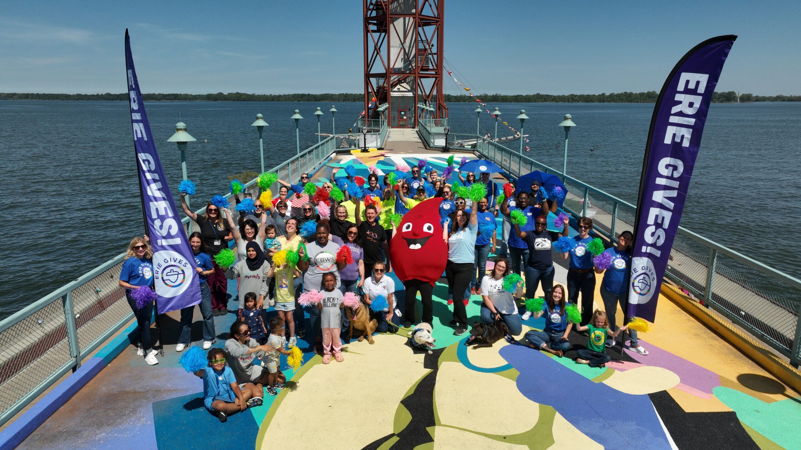A large group of people, including children and adults, poses on a colorful pier by a lake. They hold pom-poms and are joined by a red mascot. Erie Gives flags are displayed on both sides of the group.
