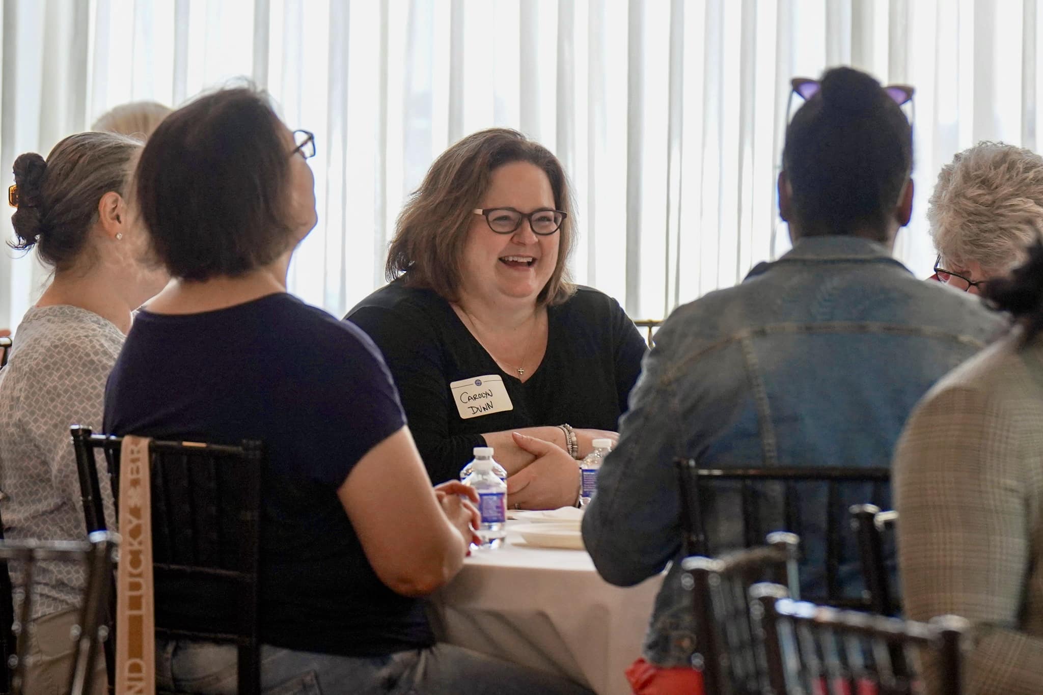 A group of people sit around a table in conversation. One woman wearing glasses and a name tag smiles while holding a pen. Bottled water and papers are on the table. Light filters through sheer curtains in the background.