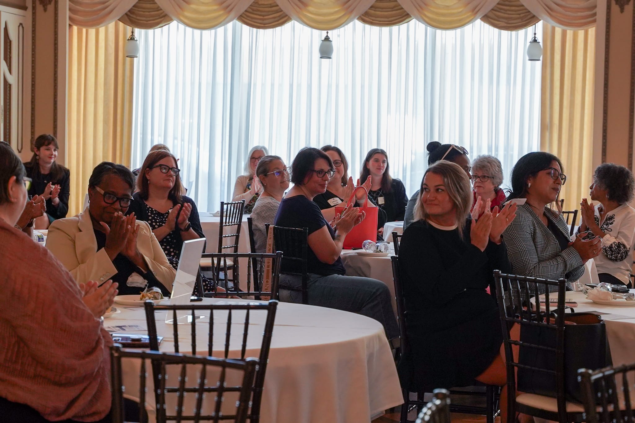 People sit at round tables and clap during an indoor daytime event. Most attendees appear to be women, engaged and looking toward the front of the room. Light streams in through large windows behind them.