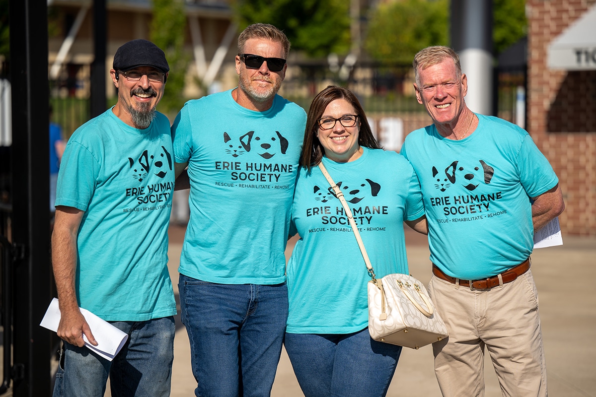 Four smiling adults stand together, all wearing matching turquoise “Erie Humane Society” T-shirts, outdoors on a sunny day. Three are men and one is a woman, who is carrying a white purse.
