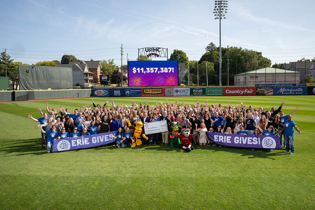 A large group of people and mascots pose on a baseball field, holding Erie Gives banners. Behind them, a scoreboard displays $11,357,387! with fireworks graphics. Its a sunny day with clear skies.