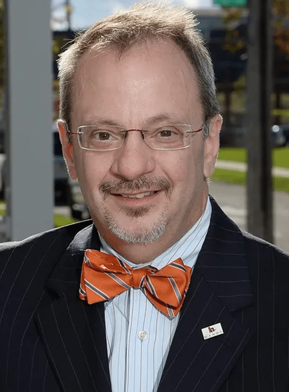 A man with glasses and a mustache smiles at the camera. He is wearing a dark pinstripe suit, a white shirt, an orange bow tie with stripes, and a pin on his lapel. The background is outdoors with blurred greenery.