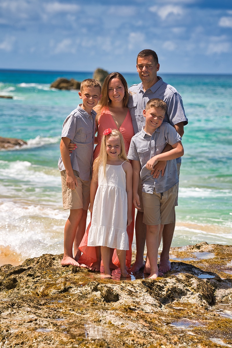 A family of five stands barefoot on rocks by the beach, smiling. The parents are behind their three children—two boys and a girl in a white dress—against a backdrop of turquoise ocean and blue sky.