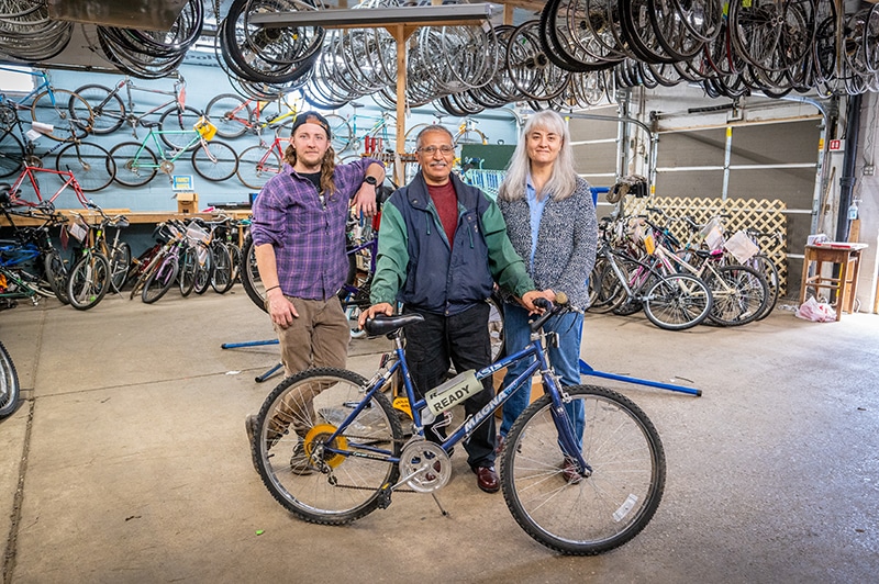 Three people stand together in a bicycle workshop, surrounded by bicycles and hanging wheels. They are posing and smiling behind a blue bike with a “READY” tag on it. The setting is spacious and well-lit.