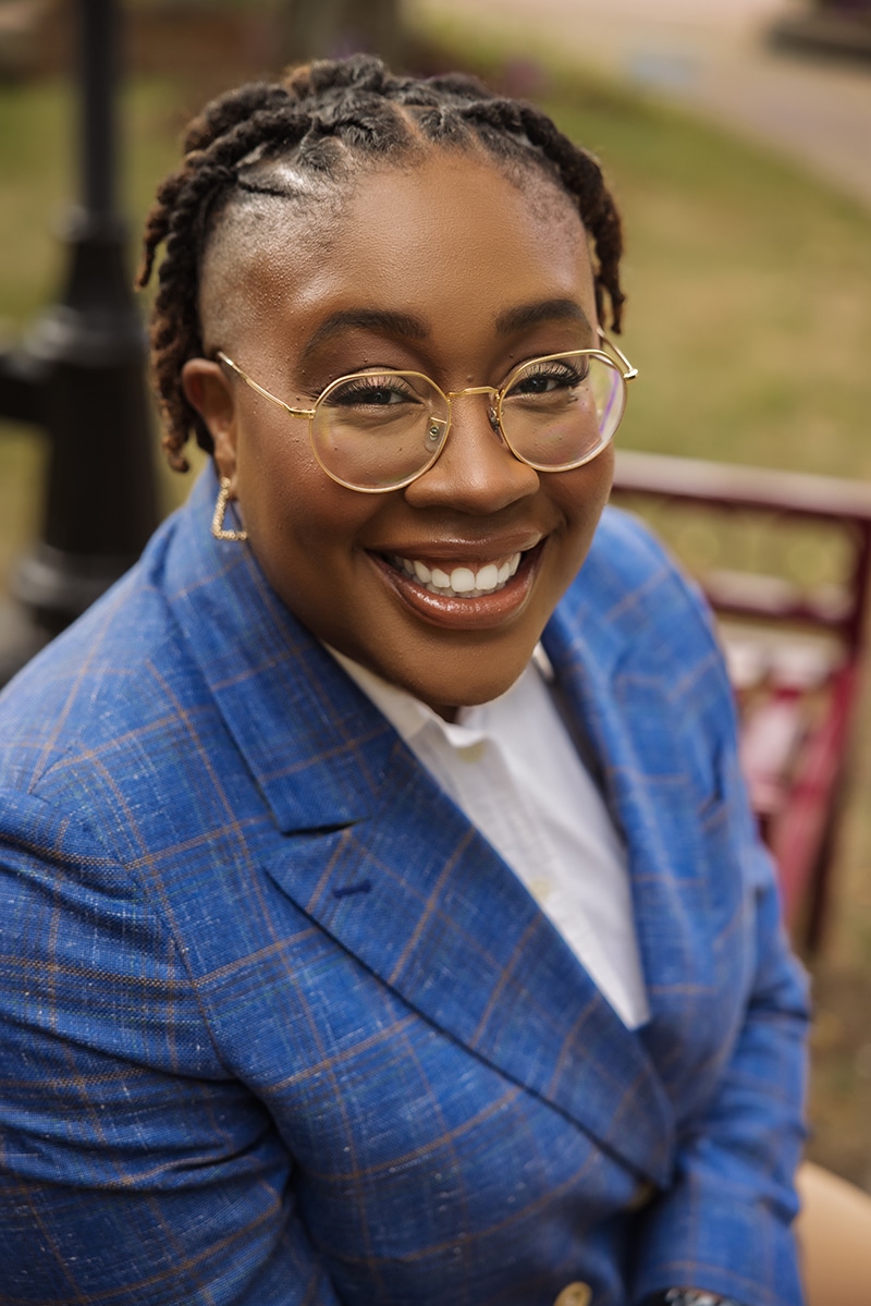 A person wearing glasses and a blue plaid blazer smiles warmly while sitting outdoors, with blurred grass and a bench in the background.