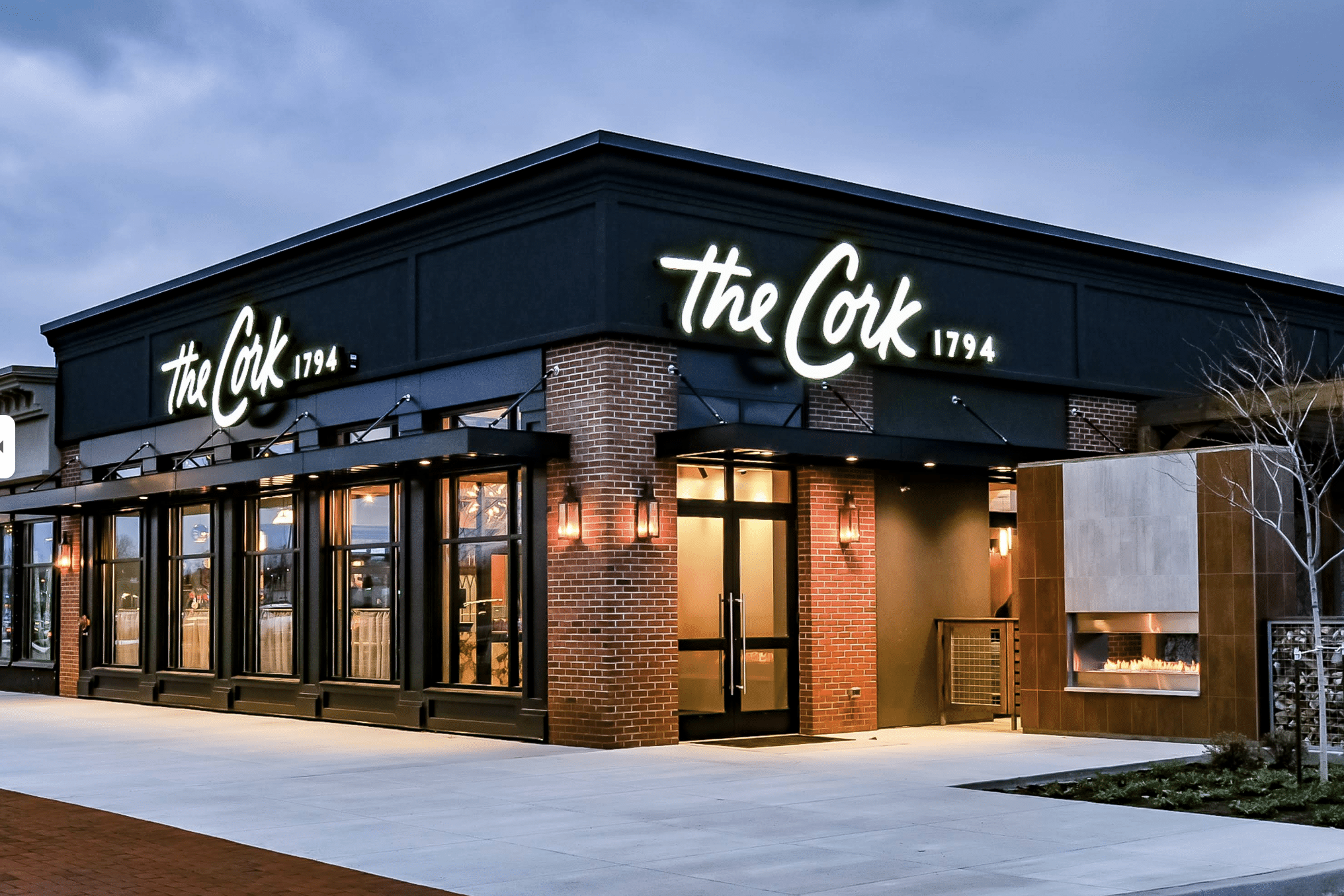 A modern restaurant exterior with large windows, brick walls, and a glowing sign reading the Cork 1794. Warm lights illuminate the entrance and a glass-enclosed fireplace sits outside on a cloudy evening.
