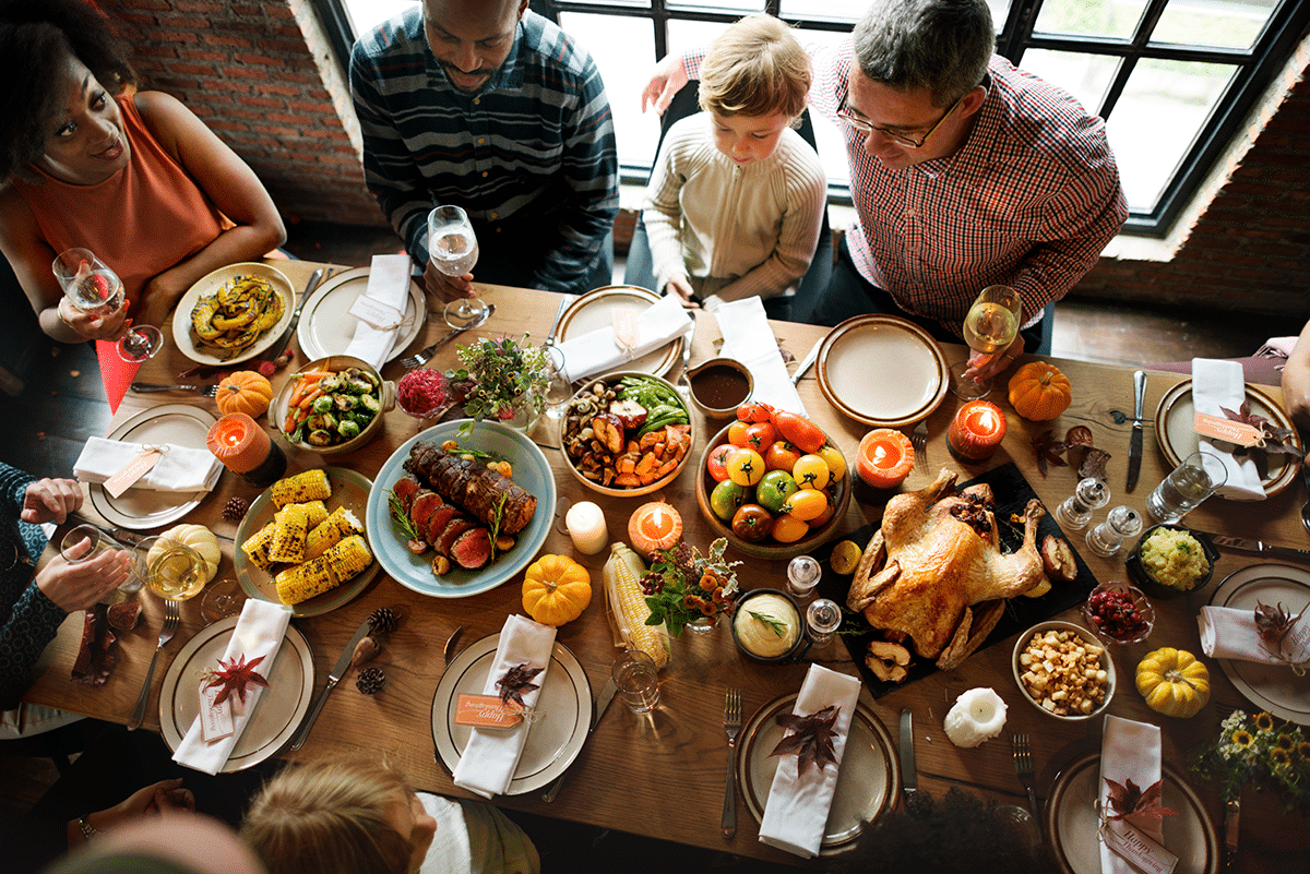 A group of people sit around a wooden table filled with various dishes, fruits, and candles, enjoying a festive meal together. The atmosphere appears warm and inviting, with a variety of colorful foods displayed.
