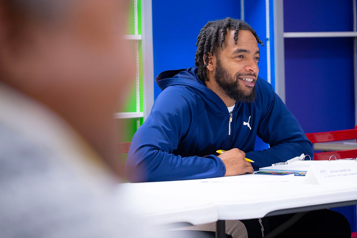 A man with braided hair and a beard, wearing a blue hoodie, sits at a table and smiles. He is holding a pencil with notebooks and papers in front of him. The foreground is blurred, focusing on the man.