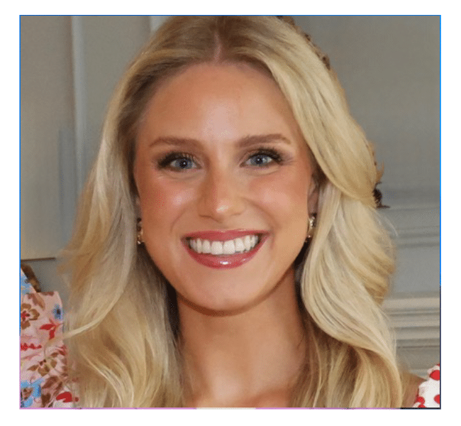 A woman with long, wavy blonde hair smiles brightly at the camera. She is wearing a floral patterned top and small hoop earrings, and is pictured indoors with a blurred background.