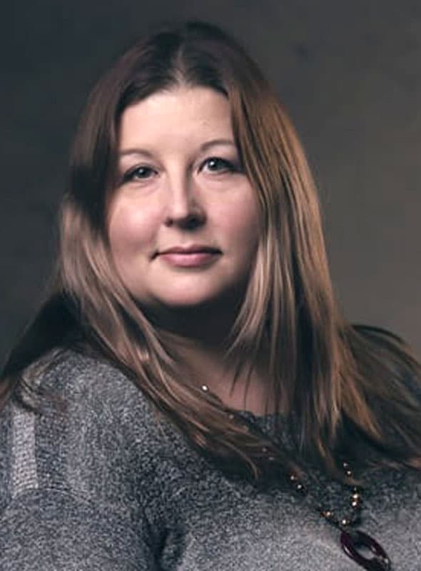 A woman with long, straight, light brown hair and fair skin wears a gray sweater and a beaded necklace, sitting in front of a plain, dark background and looking softly at the camera.