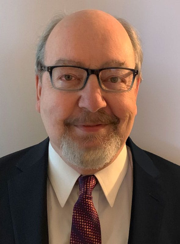 A middle-aged man with glasses, a short white beard, and thinning hair is wearing a dark suit, white shirt, and a patterned red tie, smiling slightly at the camera against a plain background.