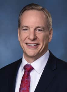 A middle-aged man with short, light brown hair smiles while wearing a dark suit, white dress shirt, and a red paisley tie against a blue background.