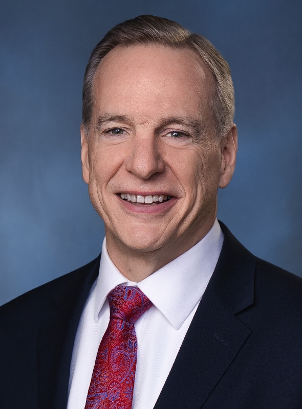 A middle-aged man with short, light brown hair smiles while wearing a dark suit, white dress shirt, and a red paisley tie against a blue background.