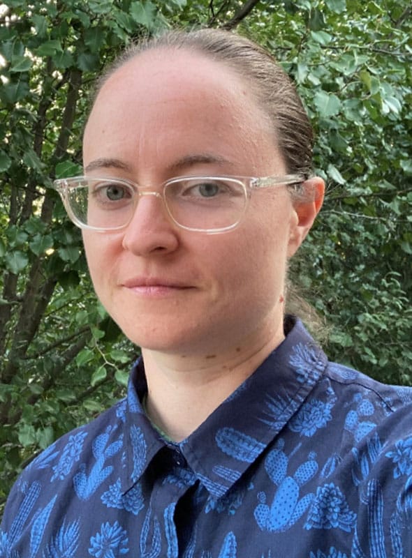 A person with light skin, glasses, and brown hair pulled back is standing outdoors in front of leafy green trees, wearing a blue shirt with a cactus pattern.