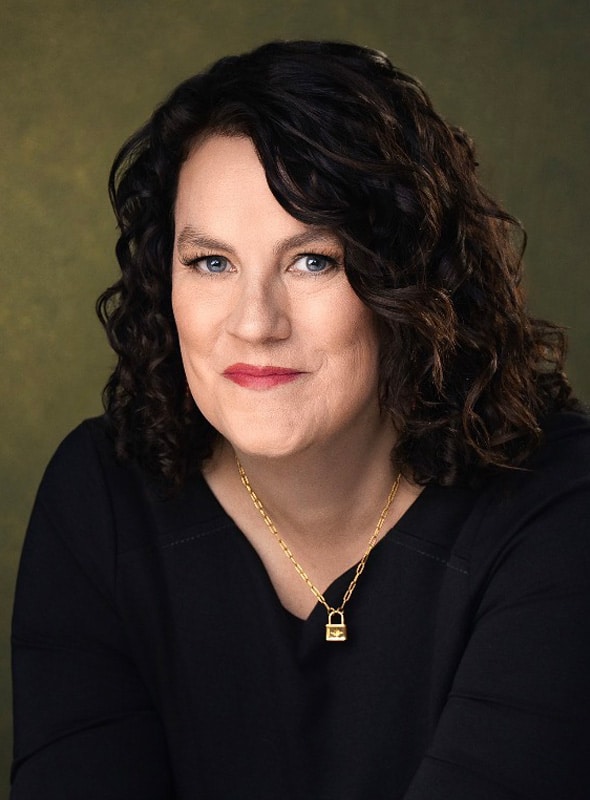 A woman with curly dark hair and blue eyes, wearing a black top and a gold necklace with a lock pendant, smiles softly against a muted green background.