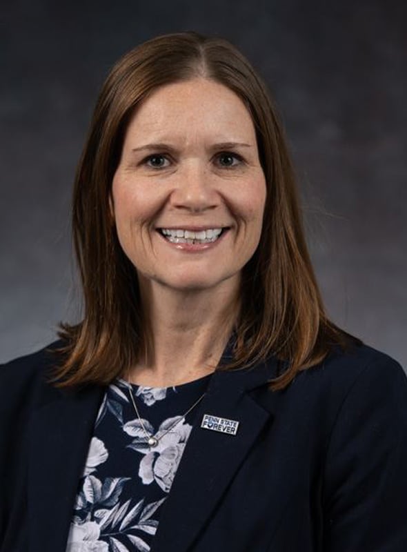 A woman with straight, shoulder-length brown hair smiles at the camera. She is wearing a dark blazer over a floral blouse and a pin that reads Penn State Forever. The background is plain and dark.