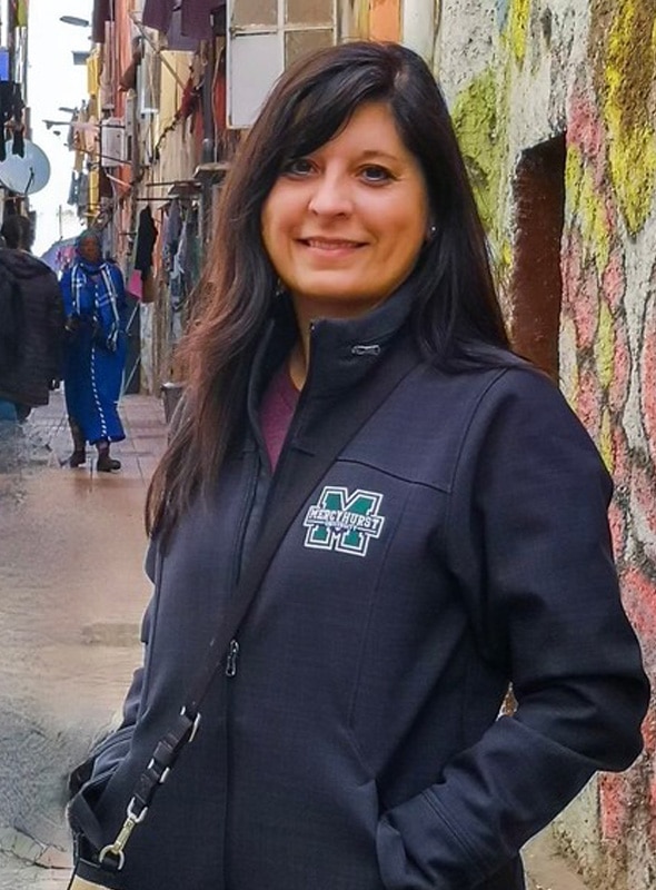 A woman with long dark hair, wearing a black Mercyhurst jacket, stands and smiles on a colorful street with painted walls and people walking in the background.