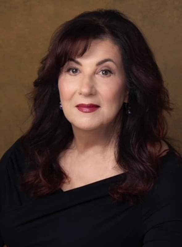 A woman with long, dark wavy hair and light skin wearing a black top poses in front of a brown background, looking at the camera with a calm expression.