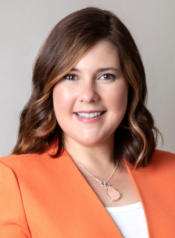 Smiling woman with shoulder-length brown hair, wearing an orange blazer over a white top and a stone pendant necklace, posing in front of a plain light background.