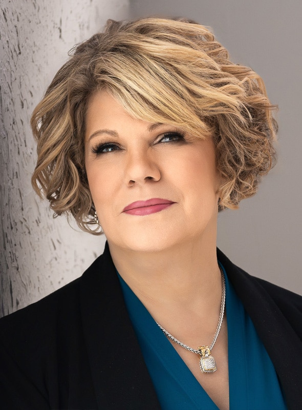 A woman with short, wavy blonde hair and light makeup wears a teal blouse, black blazer, and a silver necklace with a rectangular pendant. She looks confidently at the camera against a light-colored background.