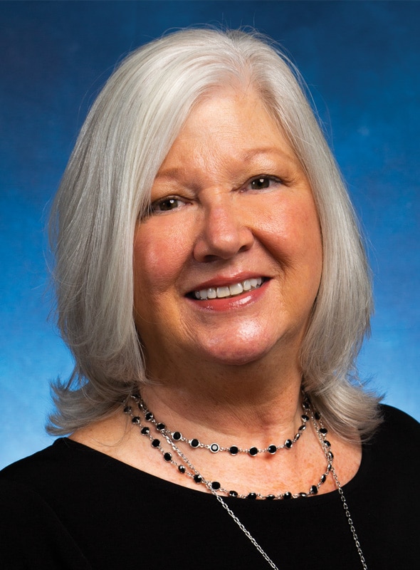 A smiling older woman with shoulder-length white hair is wearing a black top and layered necklaces, posed in front of a blue background.
