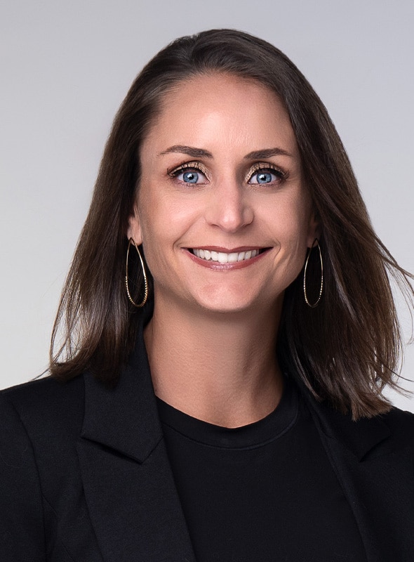 A woman with straight brown hair, blue eyes, and hoop earrings smiles at the camera. She is wearing a black blazer over a black top against a light gray background.