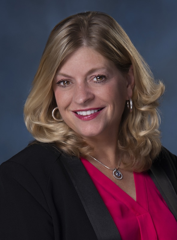 A woman with shoulder-length blonde hair smiles at the camera. She is wearing a black blazer over a bright pink blouse, hoop earrings, and a necklace with a circular pendant. The background is dark blue.