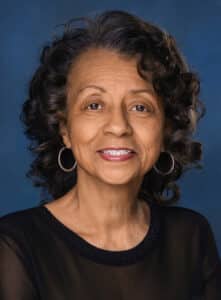 A smiling older woman with curly dark hair wearing hoop earrings and a sheer black top, posed against a blue background.