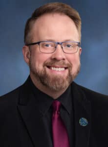A man with light skin, short brown hair, a beard, and glasses, wearing a black suit, black shirt, and dark red tie, smiling in front of a blue background.