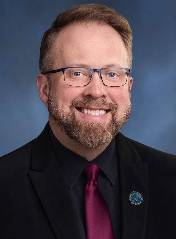 A man with light skin, short brown hair, a beard, and glasses, wearing a black suit, black shirt, and dark red tie, smiling in front of a blue background.