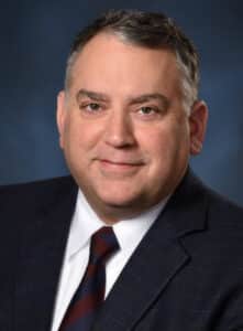 A middle-aged man with short gray hair wearing a dark plaid suit jacket, white shirt, and striped tie, smiling and posing in front of a dark blue background.