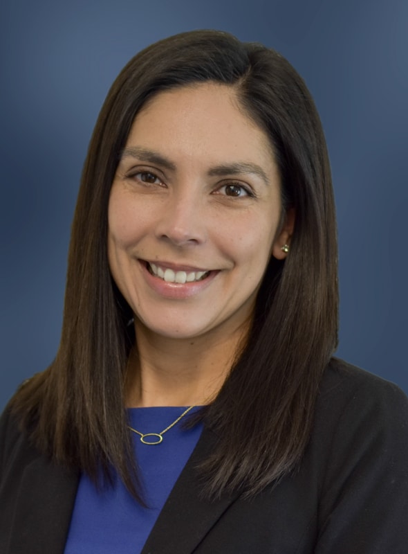 A woman with straight dark brown hair, wearing a blue top, black blazer, and a gold necklace, smiles at the camera against a solid blue background.