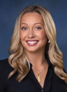 A woman with long, wavy blonde hair smiles at the camera. She is wearing a black top, gold earrings, and a gold necklace, and is posed in front of a blue background.
