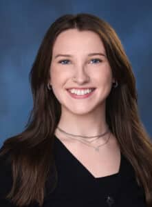 A young woman with long brown hair and blue eyes smiles at the camera. She is wearing a black top and layered silver necklaces, posed in front of a blue background.