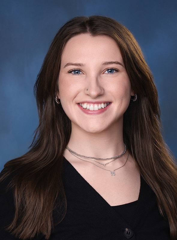 A young woman with long brown hair and blue eyes smiles at the camera. She is wearing a black top and layered silver necklaces, posed in front of a blue background.