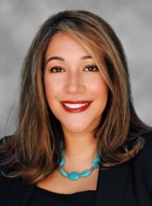 A woman with shoulder-length brown hair, wearing a black top, a turquoise beaded necklace, and a black blazer, smiling at the camera against a gray background.