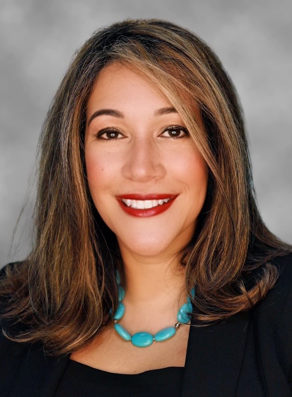 A woman with shoulder-length brown hair, wearing a black top, a turquoise beaded necklace, and a black blazer, smiling at the camera against a gray background.