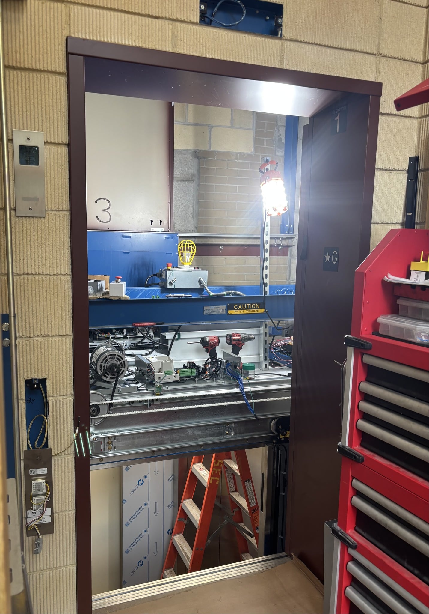 A view through an open elevator door reveals elevator repair equipment, tools, and wires. A ladder, a work light, and toolboxes are visible, with caution signs and industrial walls in the background.