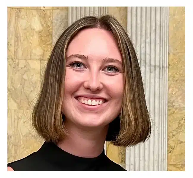 A young woman with straight, shoulder-length light brown hair smiles at the camera. She is wearing a black top and stands in front of a marble wall with a fluted column.