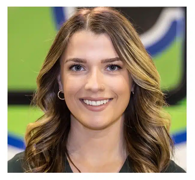 A young woman with wavy, highlighted brown hair and hoop earrings smiles at the camera. She has light skin and is posed in front of a green and blue background.