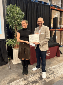 Two people stand indoors, smiling and holding a certificate together. They are in front of a potted plant and a maroon tablecloth. Shelves with cardboard boxes are visible in the background.