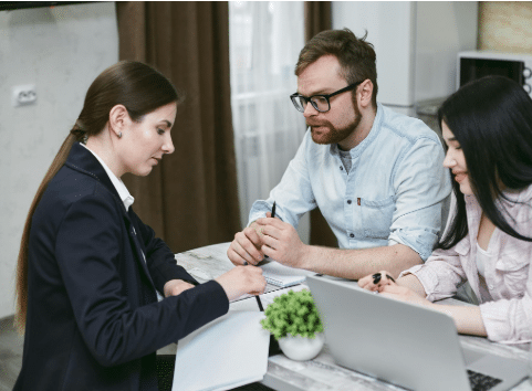 Three people sit at a table discussing documents. One woman in a suit points to papers, while a man and another woman listen attentively. A laptop and a small green plant are on the table.