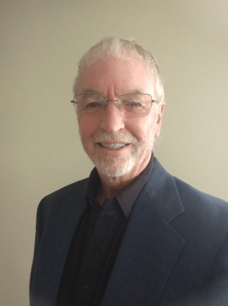 An older man with gray hair, glasses, and a short beard is smiling at the camera. He is wearing a dark jacket over a black shirt, standing against a plain light-colored background.