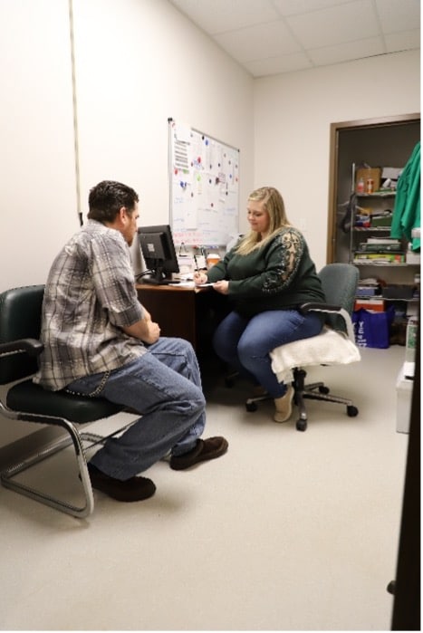 A man sits in a chair facing a woman at a desk in a small office. The woman is writing on a clipboard, and a computer and bulletin board are visible behind her. Shelves with supplies are in an open closet behind them.